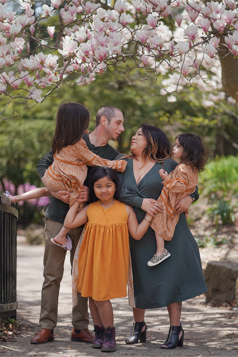 Parents in dark green clothing and their three children in orange clothing stand under a pink magnolia tree that hangs above them as they embrace